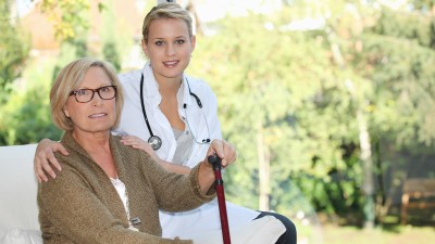 Eine ältere Frau mit einem Stock und ihre Ärztin lächeln in die Kamera.