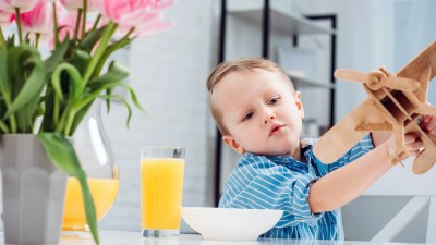 Ein kleiner Junge spielt beim Frühstück an einem Glastisch mit einem Holzflugzeug. Es steht ein volles Glas Orangensaft in seiner Landebahn.