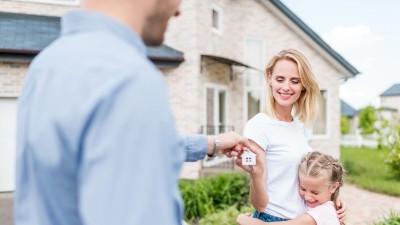 Ein Mann reicht einer Frau mit Kind einen Schlüssel mit einem Anhänger in Form eines Hauses. Sie stehen vor einem Wohngebäude.