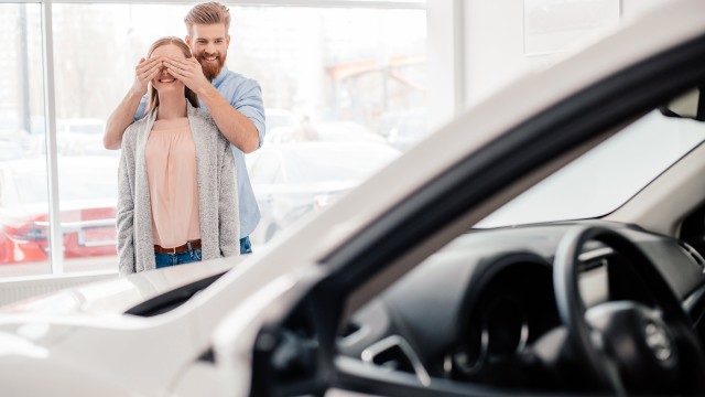 Ein Mann hält einer Frau in einem Geschäft die Augen zu, sie stehen vor einem Neuwagen.